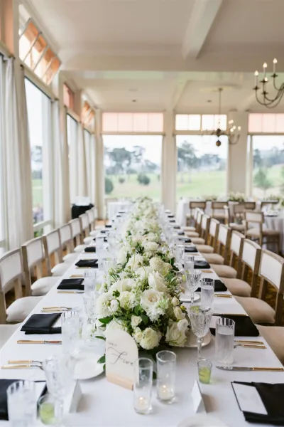 Reception tablescape with long banquet table wedding details, white floral garland and greenery, black napkins, gold flatware, and chandeliers by tall windows
