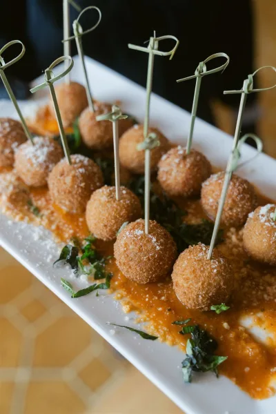 Wedding cocktail appetizers on a white platter with cocktail picks, arancini bites in marinara, topped with grated cheese and herbs on a wood table