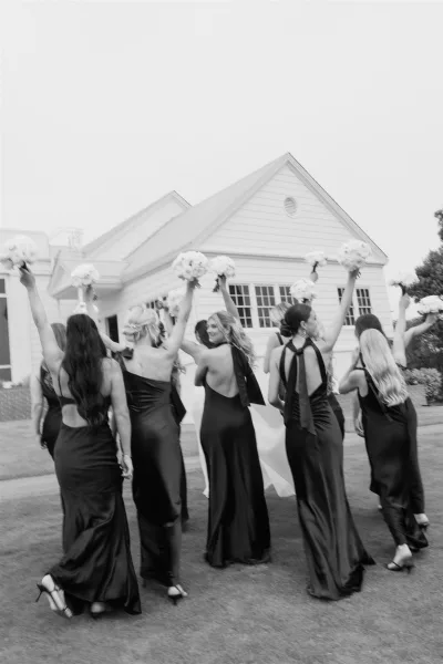 Bridesmaid group photo of bridesmaids holding bouquets in mismatched black dresses on a lawn outside a white building with tall windows