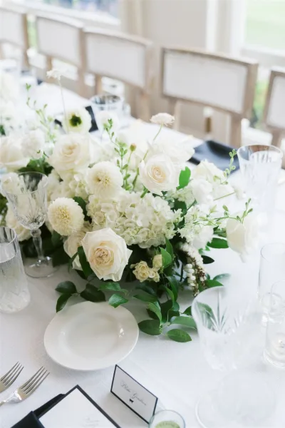 Wedding tablescape with a white floral centerpiece of roses and hydrangea, greenery garland, and candles on white linen in daylight by windows