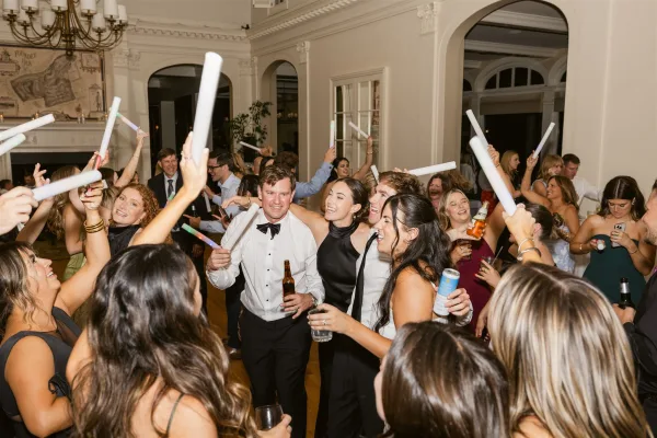 Wedding dance floor packed with reception dance party guests waving glow sticks under a chandelier in a ballroom with arched doorways and mural