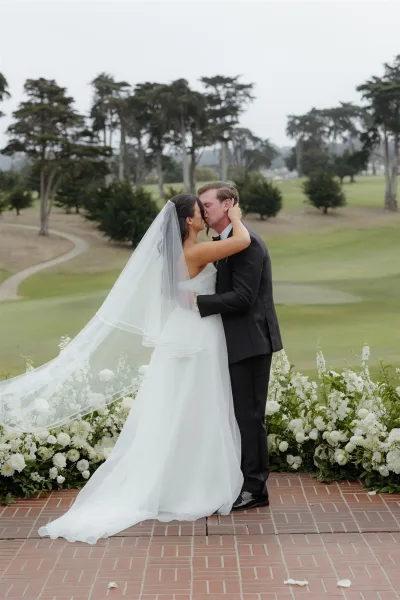 Wedding kiss portrait of bride and groom kissing, her veil blowing as they embrace on a golf course lawn beside white florals