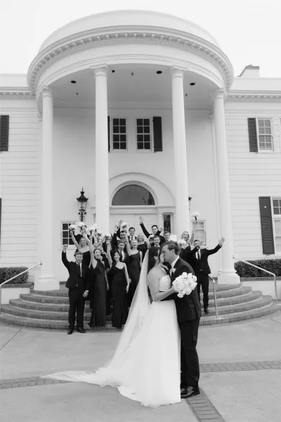 Wedding party photo as bride and groom kiss while bridesmaids and groomsmen cheer with bouquets on white columned building steps, veil flowing