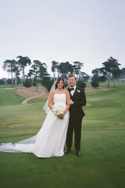 Couple portrait of bride in strapless dress with long veil holding a white bouquet beside groom in black tux on a golf course lawn