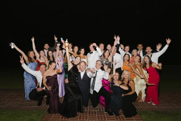 Wedding group photo of friends and family cheering with raised arms, holding beer bottles and cans on a brick patio at night