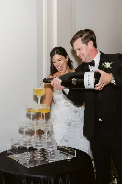 Champagne tower moment as groom pours from a bottle into stacked coupe glasses while bride in a strapless gown laughs at an indoor reception table setup