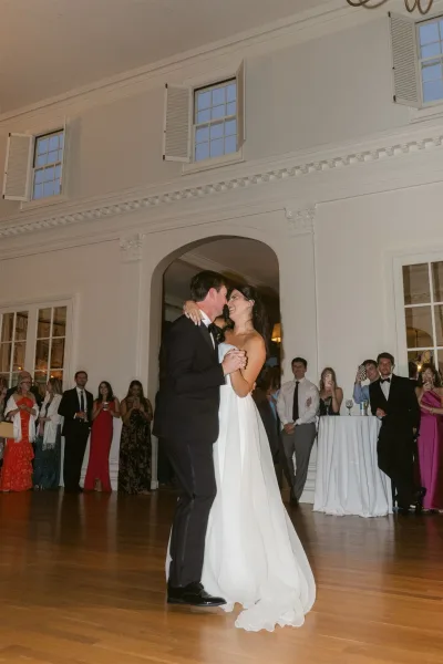 First dance as bride in a strapless wedding dress and groom in a tuxedo slow dance on a wooden ballroom floor while guests watch near cocktail tables
