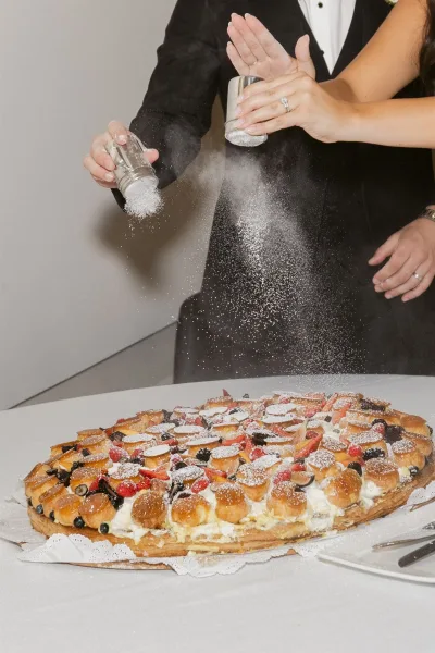 Wedding dessert fruit tart dusted with powdered sugar as bride and groom hands with rings reach in beside salt shakers on a white tablecloth