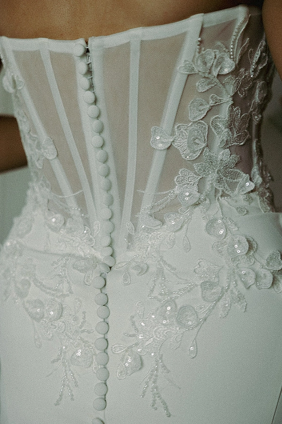 Wedding dress close-up showing a corset wedding dress bodice with lace applique, floral embroidery, sequins, and covered buttons over illusion mesh