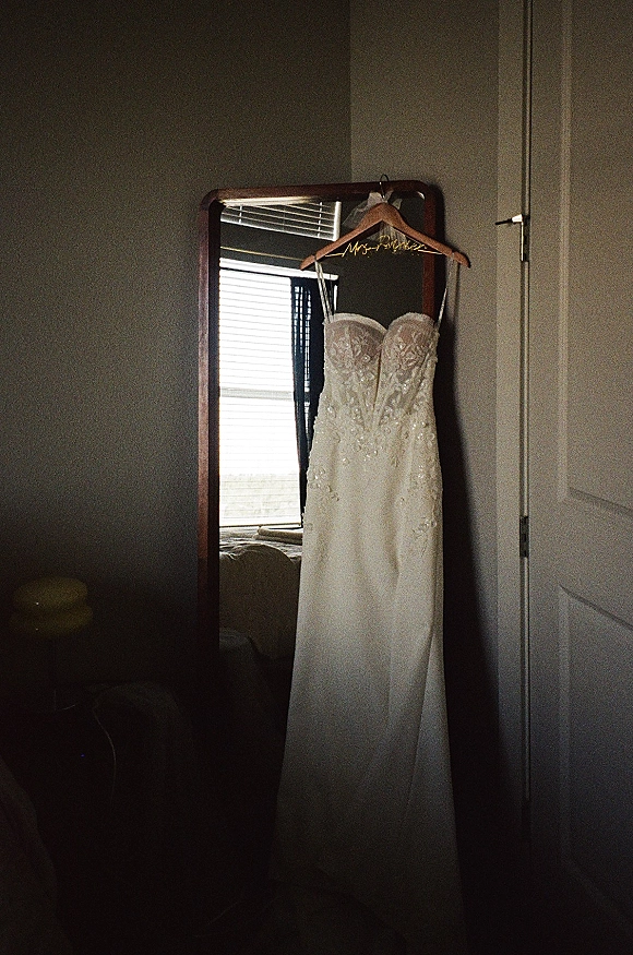 Wedding dress with lace wedding dress bodice and beading on a wooden hanger, reflected in a mirror near window blinds in a bedroom