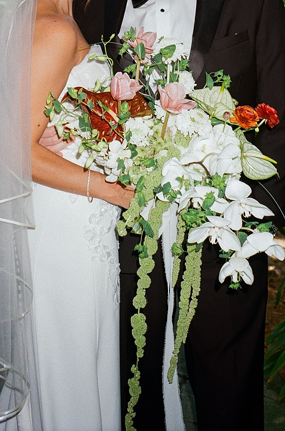 Bridal bouquet cascading with white orchids and anthurium, pink blooms, orange ranunculus, and greenery held by couple in dark indoor setting