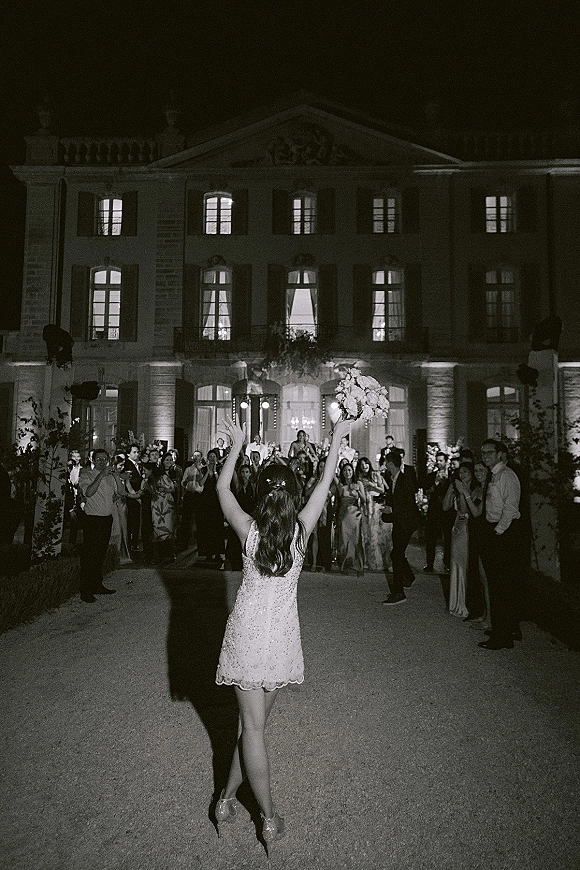 Bouquet toss at a night wedding reception send off, bride in a sparkly mini dress throws her bouquet to cheering guests in a mansion courtyard