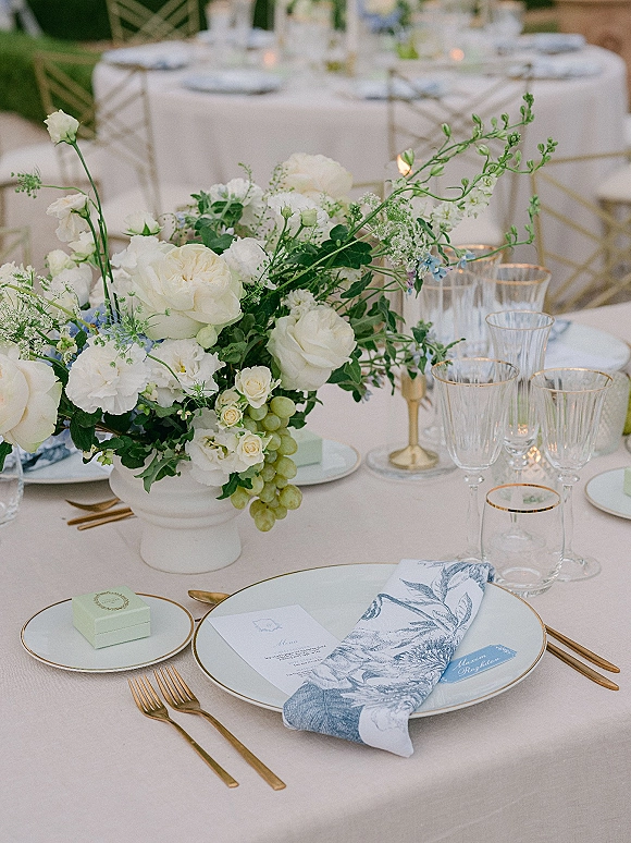 Reception tablescape with a wedding table centerpiece of white florals and greenery in a ceramic compote, gold flatware, grapes, candles on round tables