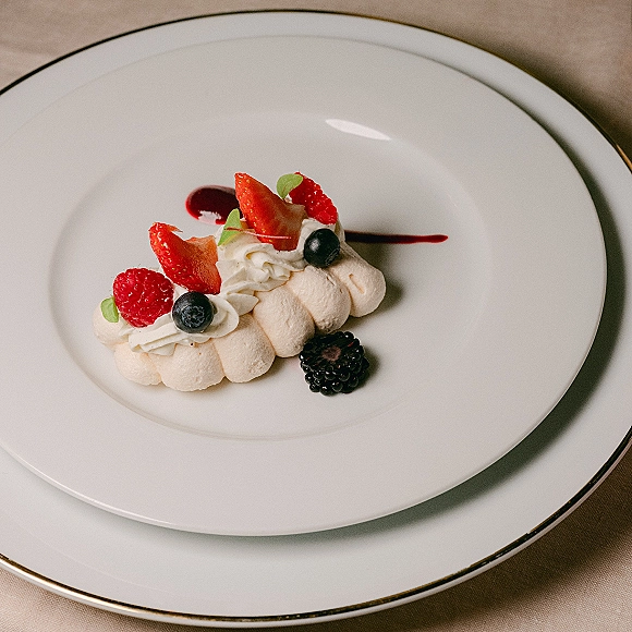 Wedding dessert plate with mini pavlova, whipped cream, and mixed berries drizzled with berry sauce on a white china gold-rim plate