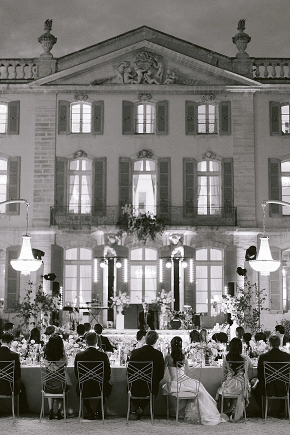 Wedding reception with a long banquet table of floral centerpieces and candles beneath chandeliers in a historic courtyard at night