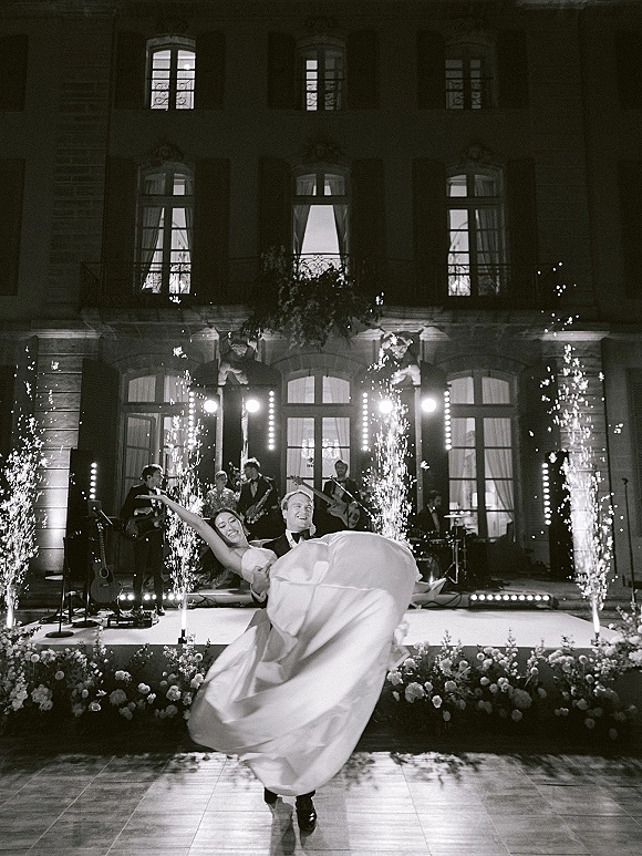 First dance moment as groom lifts bride in a wedding dance dip on an outdoor terrace dance floor with cold spark fountains and mansion backdrop at night