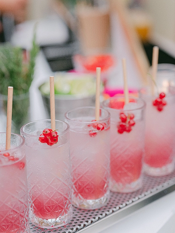 Wedding cocktails arranged on a tray in highball glasses with ice and red berry garnish at a bar counter with soft greenery behind