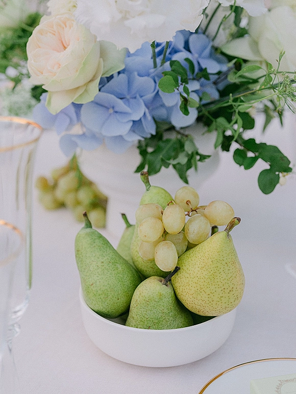 Reception tablescape with a wedding fruit centerpiece of pears and green grapes in a white bowl beside blue hydrangea and white florals on a white tablecloth