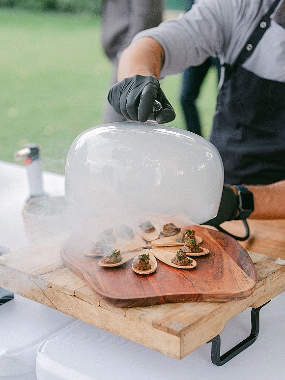 Wedding cocktail hour with passed hors d'oeuvres under a glass cloche, smoke drifting over bite-size spoons on a wooden board outdoors