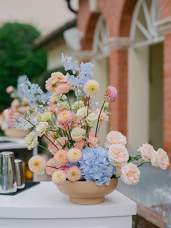 Wedding centerpiece in a low ceramic bowl with hydrangea, dahlias, roses and delphinium on an outdoor patio table by a brick building