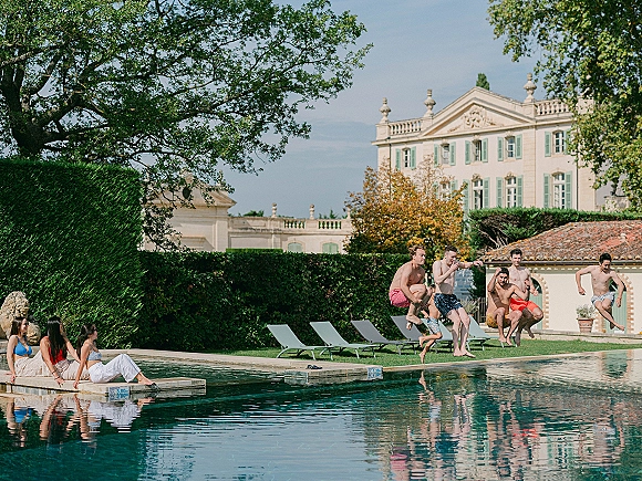 Pool party moment as friends jump into the pool in swim trunks and bikinis, splashing beside lounge chairs at a historic villa lawn setting