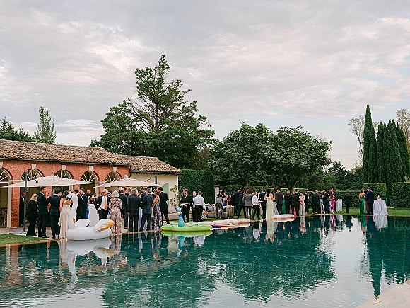 Wedding cocktail hour with guests mingling in formal attire by a pool with white umbrellas and a swan float beside a brick villa lawn