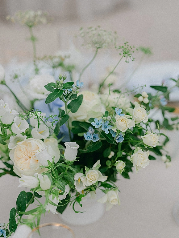 Wedding centerpiece with white flowers and blue floral centerpiece blooms in a vase with greenery on a white tablecloth at a blurred reception table