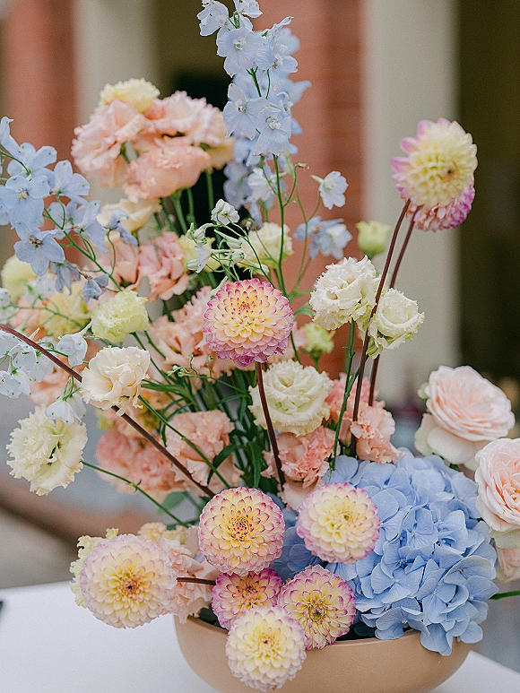 Wedding centerpiece in a ceramic bowl with pastel dahlias, hydrangea, delphinium and garden roses on a white tablecloth, venue blurred behind