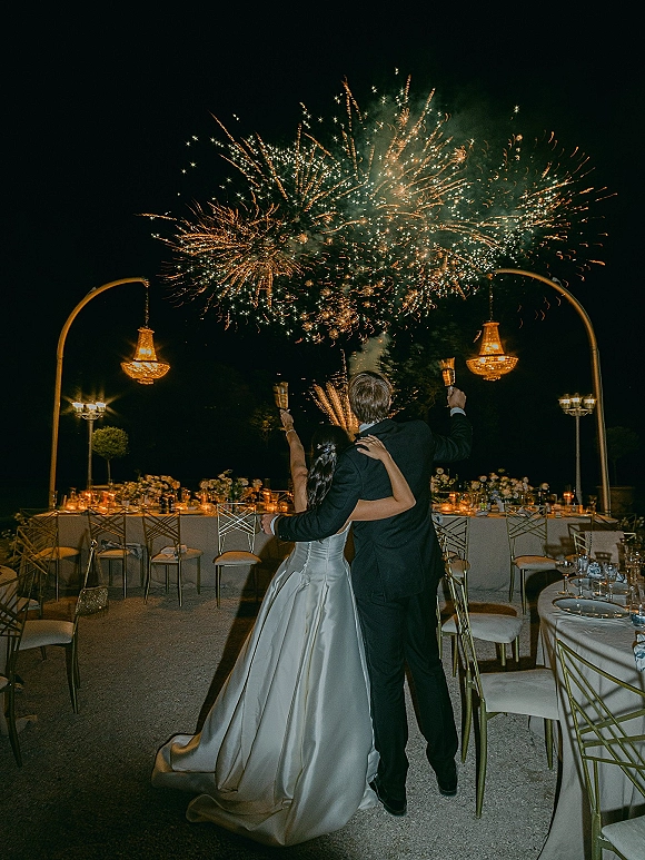 Wedding fireworks moment as bride and groom raise champagne flutes, watching bursts over candlelit terrace tables and chandeliers at night