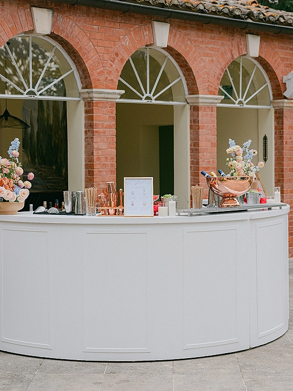 Wedding bar setup with a white counter, framed drink menu sign and copper bar tools, styled with florals in a brick-arched courtyard