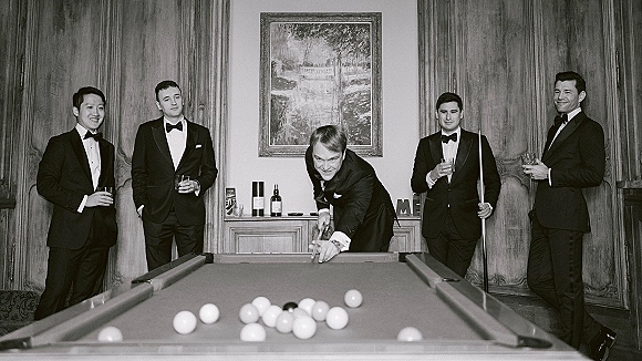 Groomsmen portrait with groomsmen playing pool in tuxedos, holding whiskey glasses around a billiards table in a wood-paneled lounge