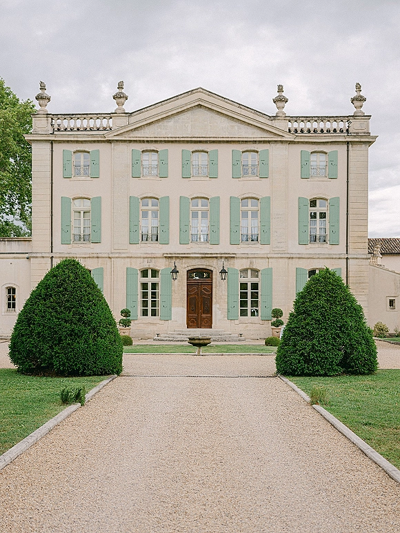 Wedding venue exterior with a stone fountain centered on a gravel driveway leading to a pale manor house entrance with trimmed hedges under cloudy sky