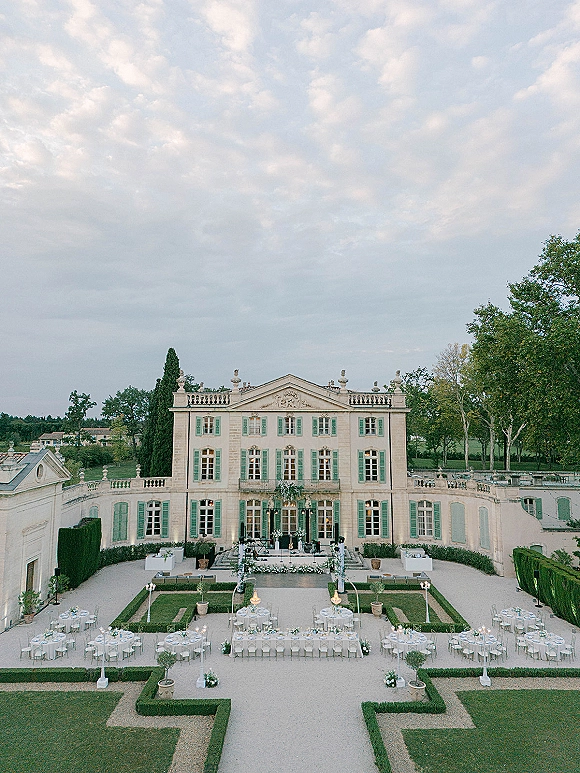 Outdoor reception setup with round tables and a long banquet table, white linens and floral centerpieces in a mansion courtyard under string lights