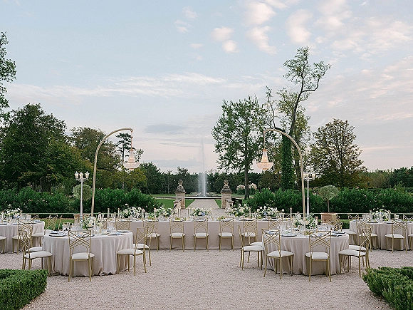 Reception tablescape with round tables and a long head table, beige linens, gold chairs, florals, and candles in a garden courtyard with fountain