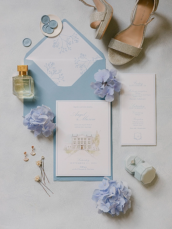 Invitation suite with blue wedding invitations, wax seal and botanical liner, styled with hydrangea, ring box, heels and perfume on white fabric