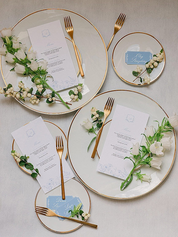 Reception tablescape with wedding place setting, gold rim plates and flatware, menu and calligraphy place cards, white florals on linen tablecloth