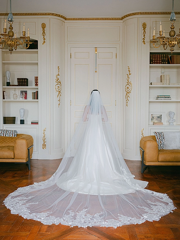 Bridal veil with cathedral length veil and lace edging drapes over wedding gown, trailing on hardwood floor in an elegant library room interior