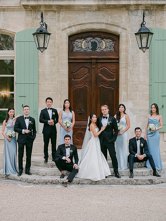 Wedding party portrait of bride and groom with wedding party on stone steps by wooden doors, bride in veil holding white bouquet