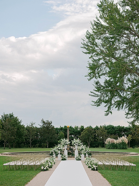 Ceremony setup with a white aisle runner leading to a white floral arch, flower-lined aisle, and chairs by a reflecting pool lawn