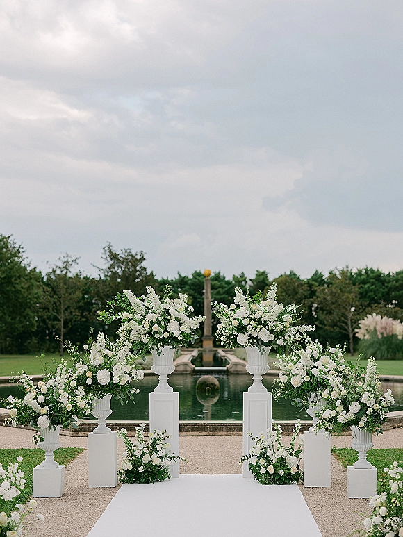 Ceremony altar with wedding ceremony backdrop of white floral arrangements and greenery along a white aisle runner by a reflecting pool