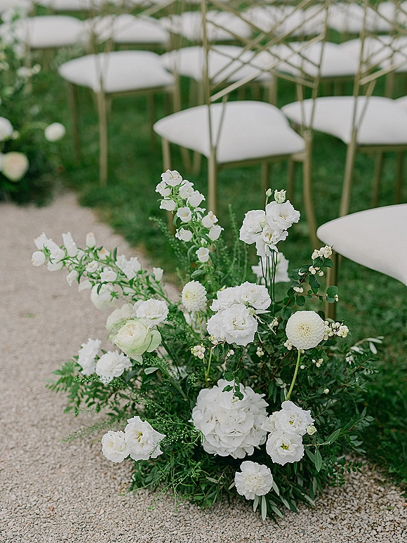 Ceremony aisle flowers and wedding aisle floral arrangements line a gravel path, with white blooms, greenery, and chairs on a lawn