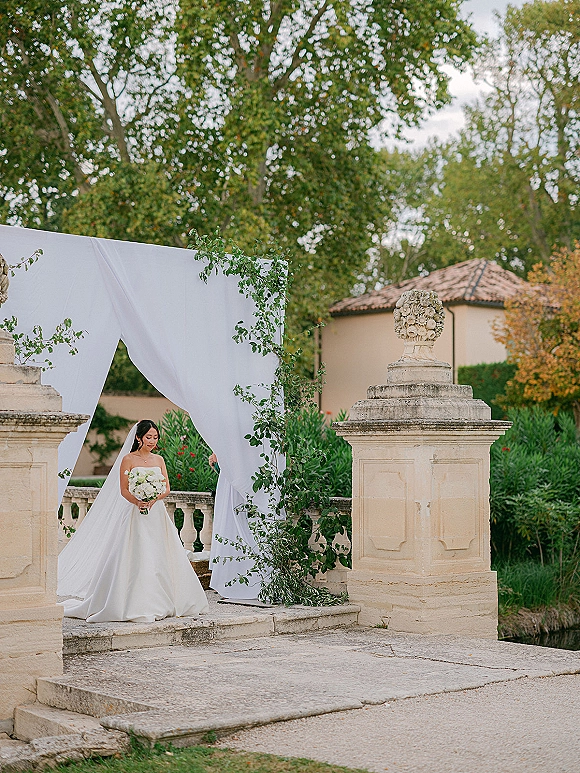 Bridal portrait of a bride holding bouquet in a strapless gown with cathedral veil, standing on a stone terrace by garden trees and pillars