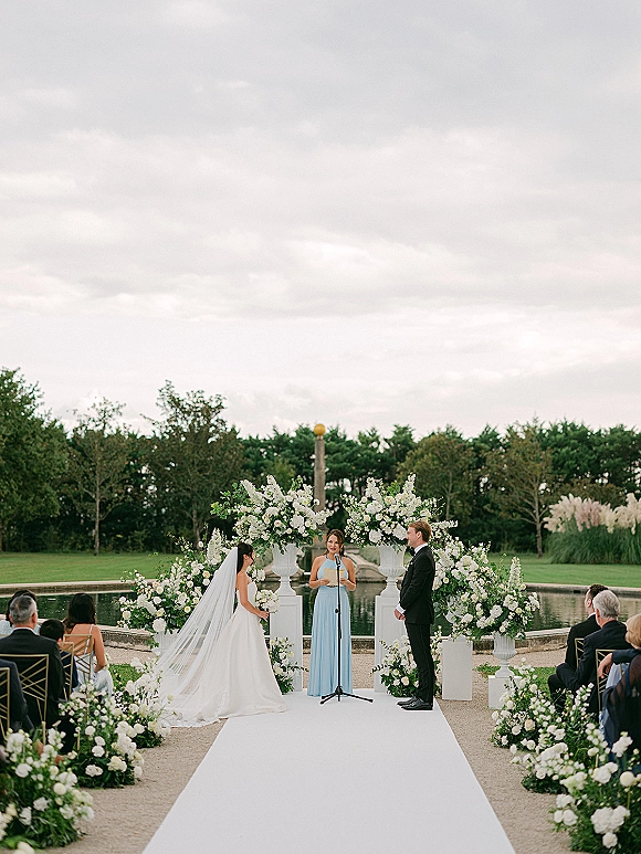 Ceremony moment at an outdoor wedding ceremony as bride and groom stand under a floral arch by a reflecting pool with white aisle runner