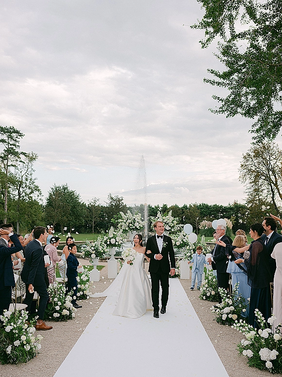 Wedding recessional as bride and groom walk the aisle on a white runner under a floral arch, petals falling with a fountain behind them