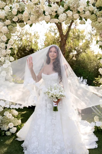 Bridal portrait of a bride with veil softly draped over her face, posing serenely in a leafy garden with trees and fresh lawn beyond.