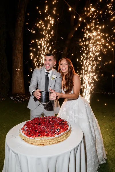 Wedding cake cutting during a candid cake cutting moment in a night garden, framed by sparkler fountains beneath a night sky.