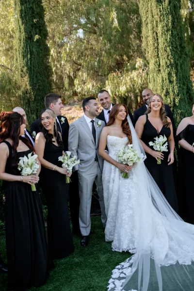 Wedding party portrait featuring the bride with bridesmaids and groomsmen, holding white calla lily bouquets amid garden cypress trees.