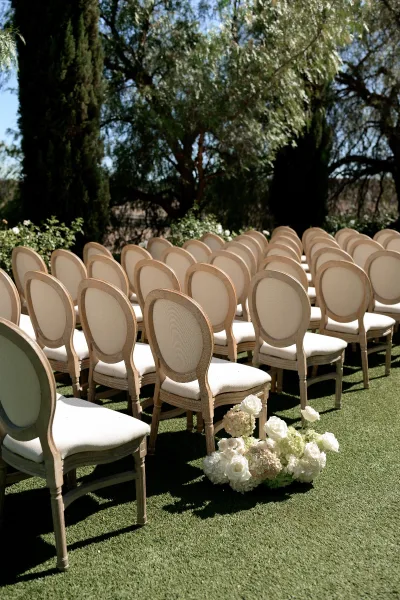 Ceremony seating in an outdoor ceremony setup lines a grass lawn, framed by white roses and hydrangeas along the aisle beneath garden trees.