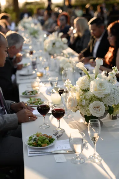 Reception tablescape on a long banquet table with a white floral centerpiece, set outdoors amid garden greenery in evening sunlight.