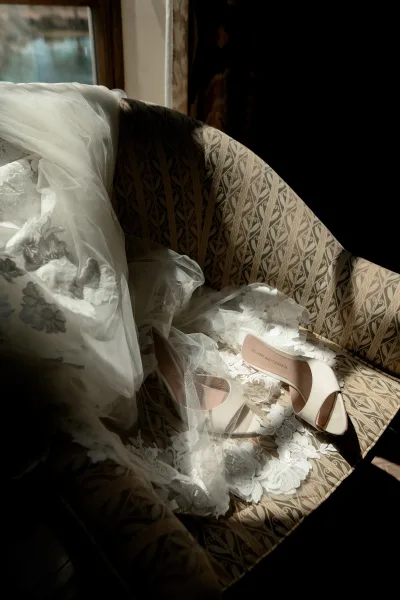 Bridal shoes, open toe bridal heels, rest on an upholstered chair, a soft veil gently draped around them.